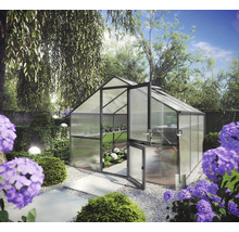 Freistehendes Gewächshaus mit grauem Rahmen und Doppelstegplatten in einem hellen Garten mit Kiesweg und violetten Blumen.