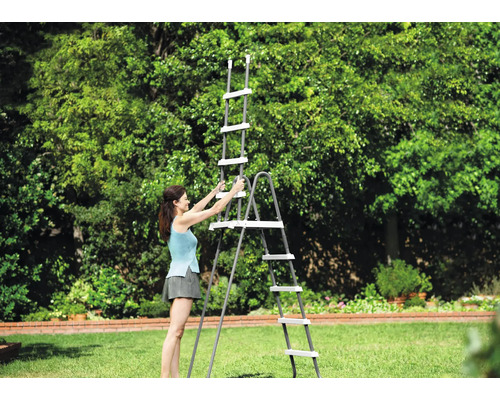 Eine Frau baut eine graue Poolleiter mit weißen Stufen auf einer Rasenfläche in einem hellen, weitläufigen Garten auf.