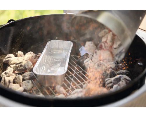 Glühende Grillbriketts werden aus einem Anzündkamin in einen Holzkohlegrill neben eine Aluminium-Tropfschale mit Weber Logo gefüllt.