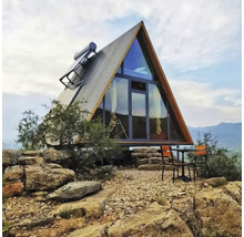 Modernes Haus in A-Form mit Glasfront, Wellblechdach und Solarpanel auf felsigem Gelände mit Tisch und Stühlen im Außenbereich.