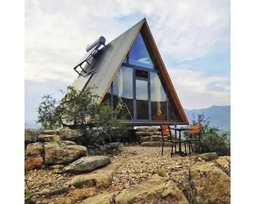 Modernes Haus in A-Form mit Glasfront, Wellblechdach und Solarpanel auf felsigem Gelände mit Tisch und Stühlen im Außenbereich.
