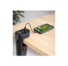 Schwarze USB-Ladestation mit Tischklemme an einer Schreibtischkante befestigt, lädt ein Smartphone über ein Kabel auf einer hellen Holzoberfläche.