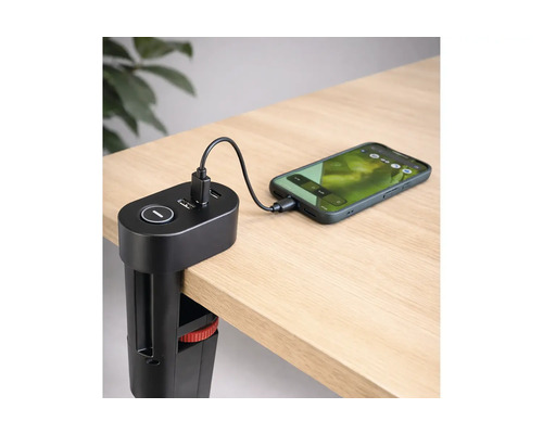 Schwarze USB-Ladestation mit Tischklemme an einer Schreibtischkante befestigt, lädt ein Smartphone über ein Kabel auf einer hellen Holzoberfläche.