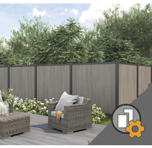Sichtschutzzaun aus WPC in einem Garten mit Holzterrasse, grauen Lounge-Möbeln aus Geflecht und blühenden Pflanzen vor Bäumen.