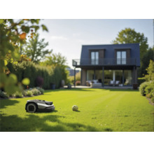 Mähroboter auf einer gepflegten Rasenfläche vor einem modernen Wohnhaus mit Terrasse und Garten.