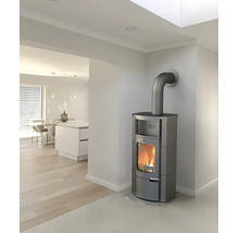 Moderner grauer Kaminofen mit brennendem Feuer in einem hellen, minimalistischen Raum mit Holzboden und offener Küche.