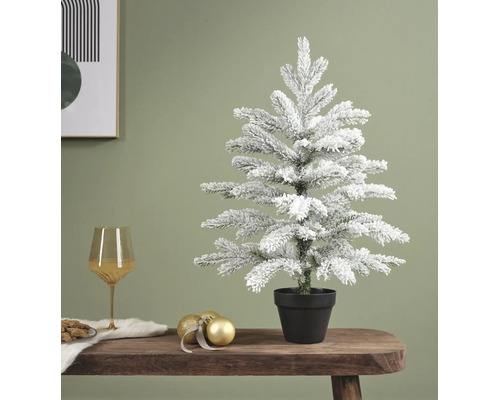Kleiner verschneiter künstlicher Christbaum im schwarzen Topf auf einem Holztisch mit goldener Weihnachtsdekoration.