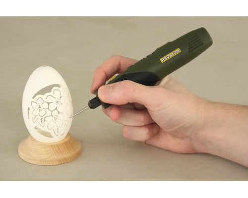 Eine Hand führt ein Proxxon Graviergerät beim Gravieren eines Blumenmusters in ein Ei auf einem Holzständer.