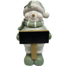 Schneemann-Figur mit Mütze und Schal, hält ein leeres schwarzes Schild an einem Holzpfosten.