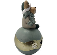 Weihnachtsfigur kleiner Bär mit Mütze und Schal sitzend auf einer dekorativen Kugel mit Schneeflockenverzierung.
