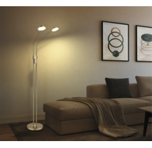 Moderne Metall-Stehlampe mit zwei verstellbaren Leuchtköpfen im Wohnzimmer neben einem Sofa mit Kissen und Wandbildern.