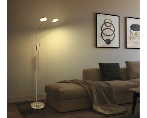 Moderne Metall-Stehlampe mit zwei verstellbaren Leuchtköpfen im Wohnzimmer neben einem Sofa mit Kissen und Wandbildern.