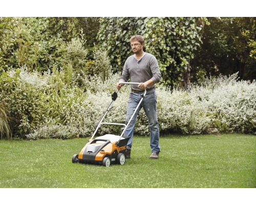 Ein Mann benutzt einen Stihl Vertikutierer auf einer Rasenfläche in einem Garten mit Sträuchern.