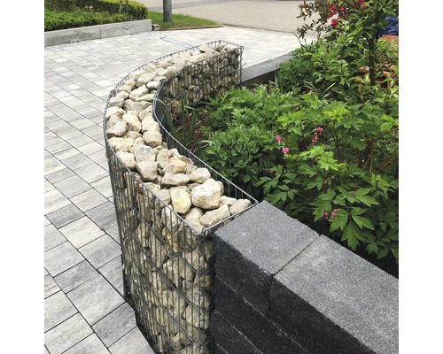 Gebogene Gabione mit Steinfüllung als Gartenbegrenzung neben einer Mauer und Pflanzen auf gepflastertem Untergrund.
