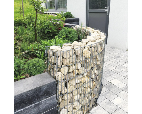 Geschwungene Gabione aus Drahtgeflecht mit Naturstein-Füllung als Beetumrandung im Garten neben einer Pflasterfläche.