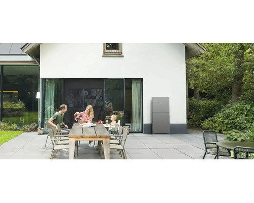 Familie auf Terrasse mit großem Holztisch vor einem Haus mit installiertem grauem Batteriespeicher an der Außenwand.