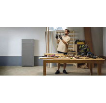 Werkstatt mit einem Mann an einer Holzwerkbank, Handwerkzeugen und einem grauen Heimspeichersystem an der Wand in heller Atmosphäre.