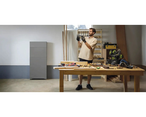 Werkstatt mit einem Mann an einer Holzwerkbank, Handwerkzeugen und einem grauen Heimspeichersystem an der Wand in heller Atmosphäre.