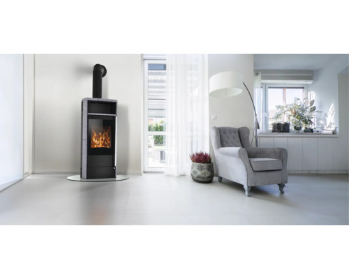 Helles, modernes Wohnzimmer mit einem schwarzen Kaminofen mit sichtbarem Feuer, einem grauen Sessel und einer Glastür nach draußen.