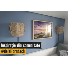 Blaue Wand dekoriert mit einem Holzbilderrahmen mit Landschaftsmotiv und zwei Wandlampen aus Naturgeflecht über einem weißen Bett.