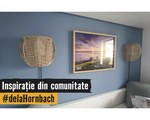 Blaue Wand dekoriert mit einem Holzbilderrahmen mit Landschaftsmotiv und zwei Wandlampen aus Naturgeflecht über einem weißen Bett.