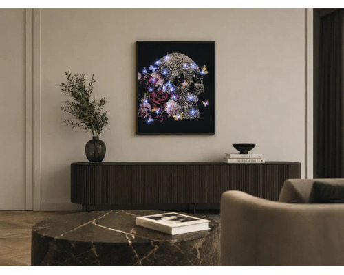Ein gerahmtes Wandbild mit einem Totenkopf, Blumen, Schmetterlingen und blauen LED-Lichtern über einem dunklen Sideboard in einem modernen Wohnzimmer.