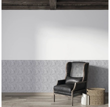 Helle Wandgestaltung mit Vliestapete in Steinoptik unten und weißer Wand darüber. Schwarzer Leder-Ohrensessel auf Parkett unter Holzbalkendecke.