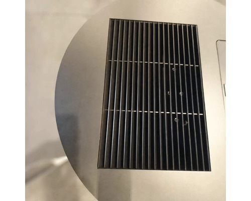 Nahaufnahme eines rechteckigen schwarzen Lüftungsgitters aus Metall, das in eine kreisförmige Edelstahloberfläche eingelassen ist.