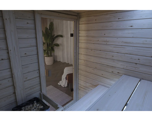 Innenansicht einer Holzsauna mit Blick durch eine Glastür in einen angrenzenden Raum mit Zimmerpflanze und Ruheliege.
