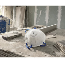 Weiße Rautner Feinstaubmaske mit Ausatemventil und blauen Kopfbändern auf einer staubigen Baustelle.