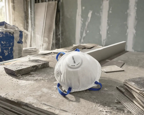 Weiße Rautner Feinstaubmaske mit Ausatemventil und blauen Kopfbändern auf einer staubigen Baustelle.