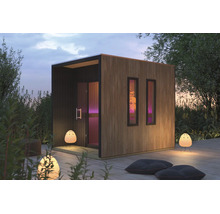 Moderne Gartensauna aus Holz mit Flachdach und Lamellenverkleidung auf einer Holzterrasse im Garten bei Dämmerung.