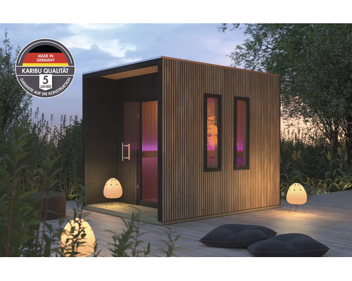 Moderne Karibu Gartensauna mit Holzlamellen Fassade und zwei Fenstern, aufgestellt auf einer Holzterrasse mit Außenbeleuchtung und Sitzkissen.