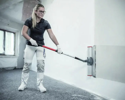 Eine Frau verwendet einen breiten Flächenspachtel mit Teleskopstange zum Glätten einer Wand in einem hellen Innenraum.