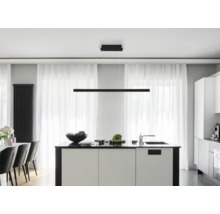 Lineare schwarze LED-Pendelleuchte über einer weißen Kücheninsel in einer modernen, hellen Küche mit großen Fenstern und transparenten Vorhängen.