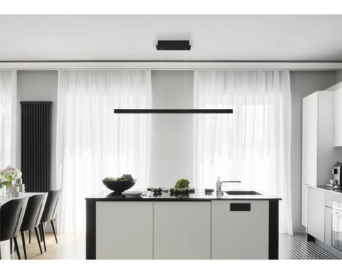 Lineare schwarze LED-Pendelleuchte über einer weißen Kücheninsel in einer modernen, hellen Küche mit großen Fenstern und transparenten Vorhängen.