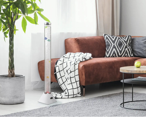 LED-Wassersäule mit dekorativen Fischen in einem hellen Wohnzimmer neben einem braunen Sofa und einer großen Zimmerpflanze.