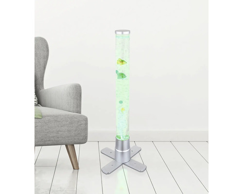 Beleuchtete Wassersäule mit dekorativen Fischen, silberfarbenem Standfuß und Luftblasen neben einem grauen Sessel auf hellem Holzboden.