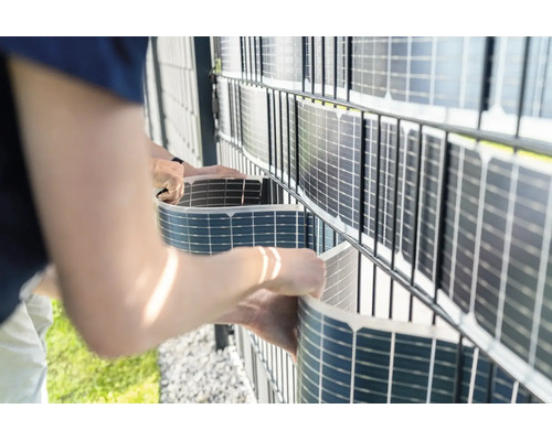 Person montiert flexible Solarmodule als Sichtschutz an einem Doppelstabmattenzaun im Garten.