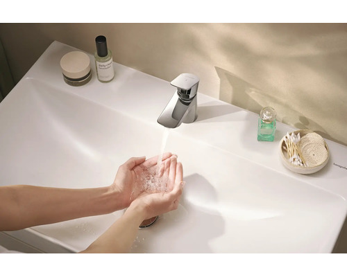 Hände waschen an einer modernen Hansgrohe Waschtischarmatur über einem weißen Waschbecken mit Kosmetikartikeln im Badezimmer