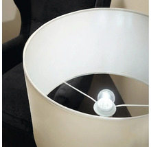 Eine elegante Stehlampe mit zylindrischem Lampenschirm und leuchtendem Leuchtmittel vor einem dunklen Sessel.