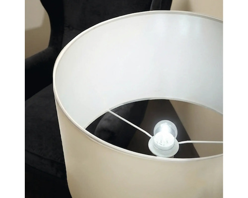 Eine elegante Stehlampe mit zylindrischem Lampenschirm und leuchtendem Leuchtmittel vor einem dunklen Sessel.