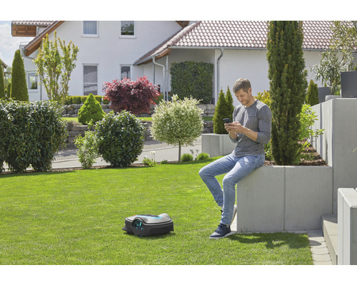 Mann sitzt im Garten und bedient Rasenmähroboter per Smartphone.