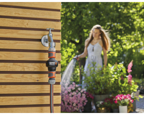 Gartenwasserzähler am Wasserhahn mit Frau im Hintergrund, die den Garten bewässert