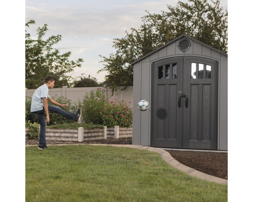 Szene mit einem Jungen, der im Garten einen Ball gegen ein graues Gartenhaus tritt