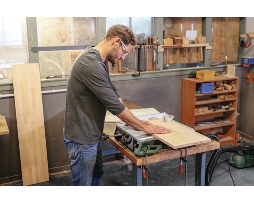 Ein Mann mit Schutzbrille arbeitet mit einer Tischkreissäge an einem Holzbrett in einer Werkstatt.