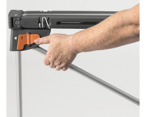 Eine Hand betätigt den orangefarbenen Verriegelungsmechanismus eines grauen Arbeitstischgestells aus Metall.