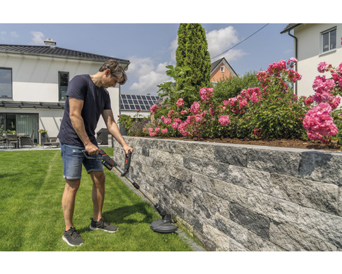 Mann schneidet Rasen mit einem Rasentrimmer an einer Steinmauer in einem Garten.