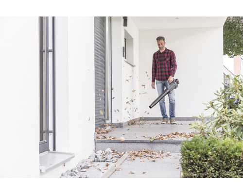 Ein Mann benutzt einen Laubbläser vor einem Haus, um Laub zu entfernen.