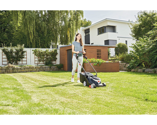 Frau bedient Rasenmäher im Garten vor Wohnhaus und Gartenhaus